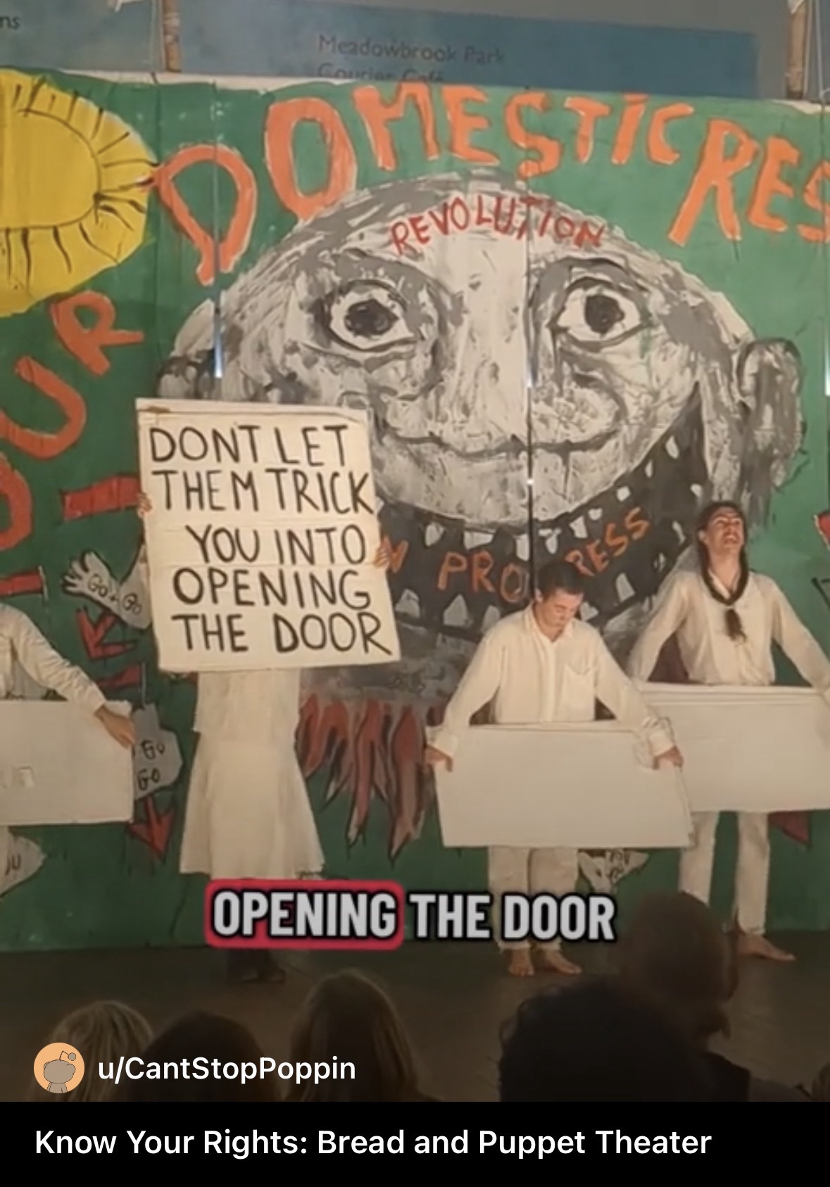 A screenshot of the bread and puppet theatre performance. They hold a sign that says “don’t let them trick you into opening the door.”
