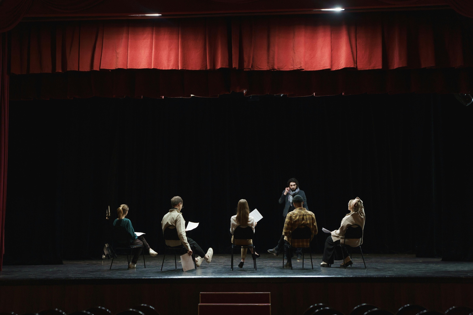 A stock image of a group of artists sitting on chairs on a stage, scripts in hand. 