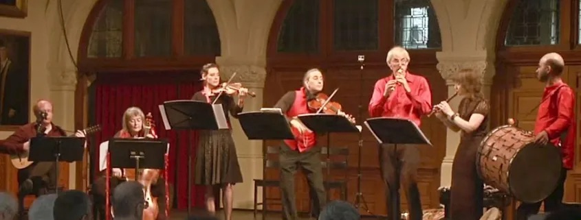A group of musicians are in a line, some are sitting with their instruments, some are standing. Some are wearing red tops. They are looking at one another with warm faces. 