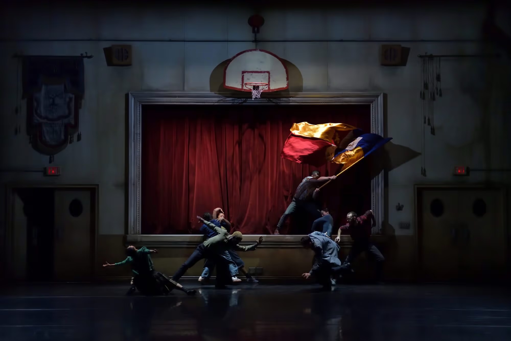A dramatically lit still from the dance show. What appears to be a school gym with a raised stage and a basketball hoop, the dancers stand in dramatic poses and unfurl an unknown flag. 